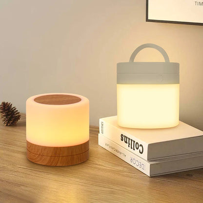 Luminária para quarto Aura 360 com base amadeirada e base branca emitindo luz quente suave em cima de uma mesa com livros.