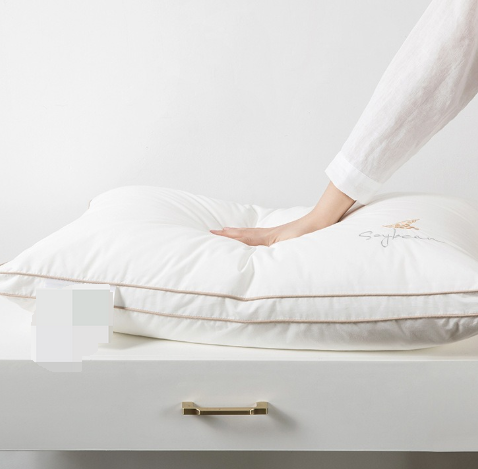 Travesseiro ortopédico Sillencia SoySoft branco com bordado Soybean e detalhes em linha dourada com uma mão mostrando a qualidade do produto.