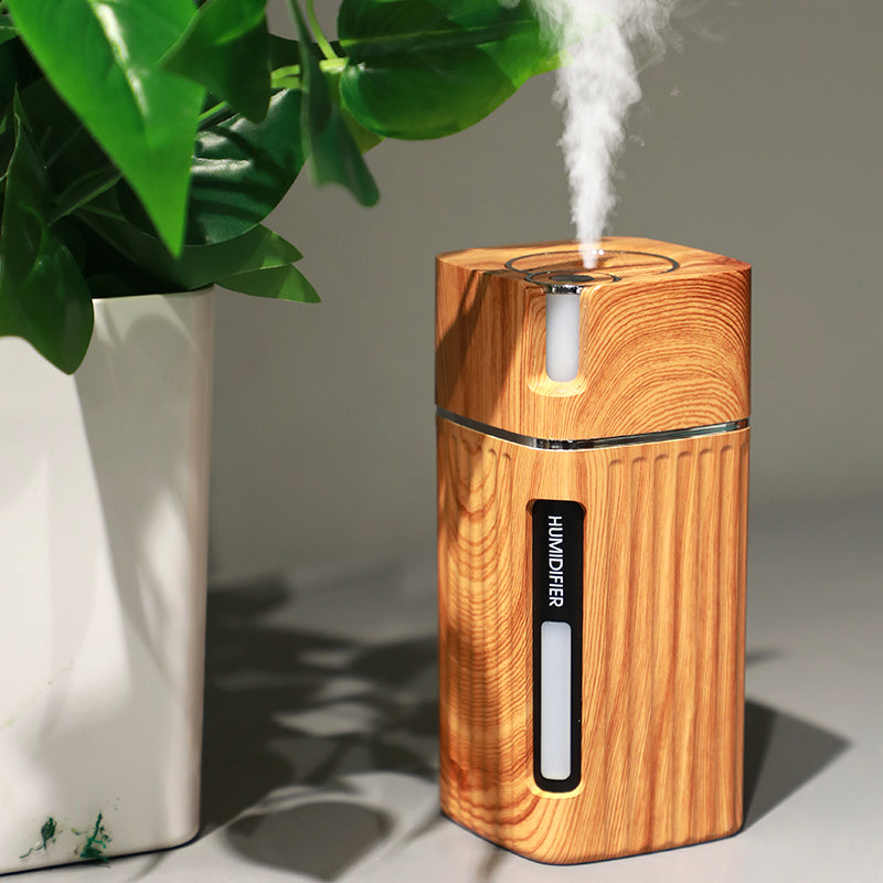 Umidificador de ar Sillencia WoodMist em textura amadeirada clara liberando névoa fina ao lado de uma planta.